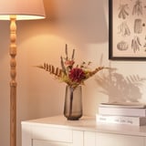 Un elegante ramo de flores en un jarrón de cristal se encuentra sobre una cómoda blanca, iluminada por una lámpara de pie de madera, añadiendo color y elegancia a la habitación.