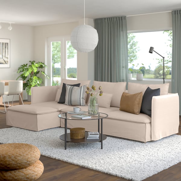 Sofá modular SÖDERHAMN beige con almohadas de colores, mesa de centro de cristal, otomana tejida, grandes ventanales con cortinas de color verde grisáceo.