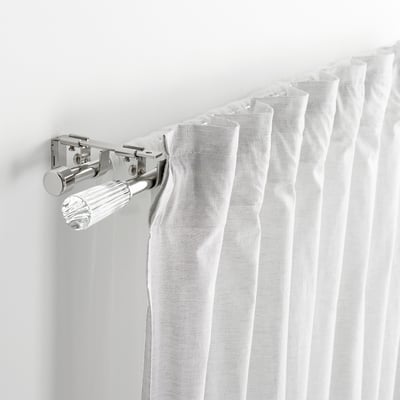 Primer plano del soporte cromado de la barra de la cortina que sostiene las cortinas blancas, mostrando parte del conjunto soldvärm.