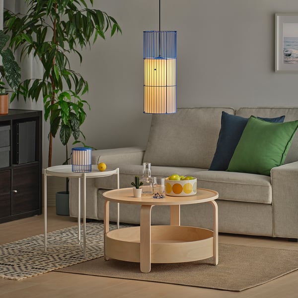 Sala de estar moderna con sofá beige, almohadas de colores y una lámpara colgante que proyecta luz decorativa. Una mesa de centro contiene fruta y un jarrón.