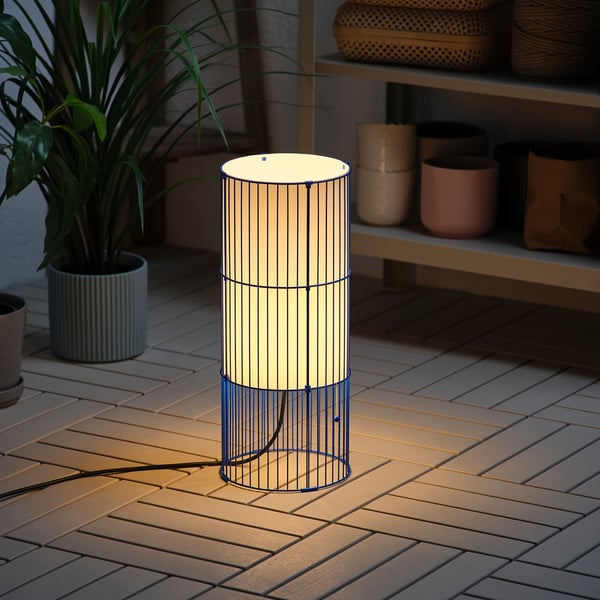 Una lámpara cilíndrica de exterior, SOLVINDEN, que da un brillo cálido sobre una cubierta de madera. Es de pie con una luz decorativa.