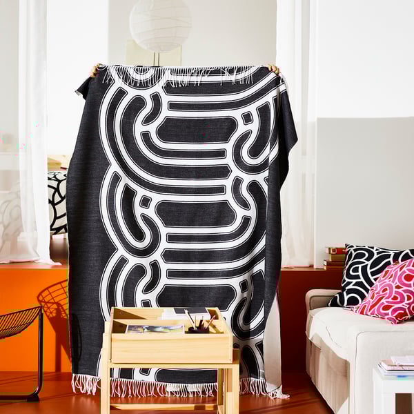 Una persona sostiene una manta abstracta en blanco y negro en una habitación con una silla de madera, un sofá con almohadas y una lámpara.