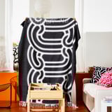 Una persona sostiene una manta abstracta en blanco y negro en una habitación con una silla de madera, un sofá con almohadas y una lámpara.