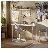 Moderno escritorio blanco con patas ajustables, con una silla ergonómica y varias decoraciones artísticas en una pared de tonos neutros.