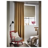 Acogedor rincón de dormitorio con sillón rojo, cojín a cuadros, lámpara de madera, cortinas beige, rollos blancos y ropa de cama suave.