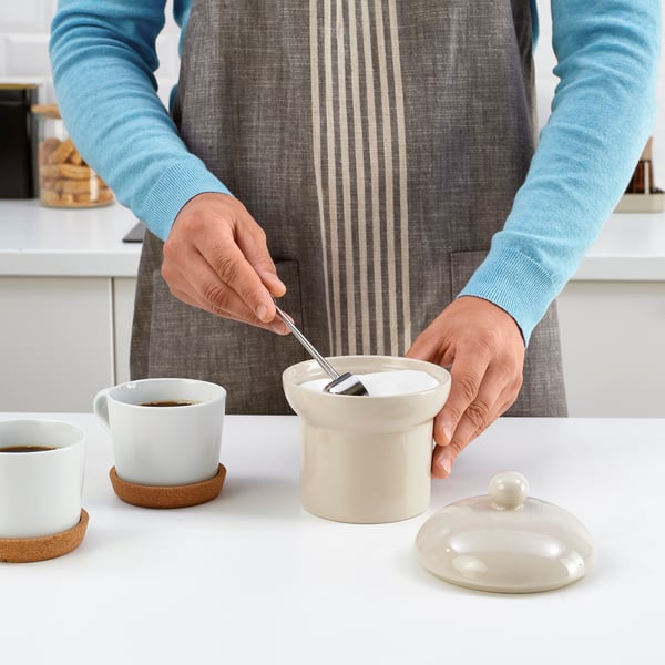 Una persona está usando una cuchara para sacar azúcar de un recipiente etiquetado como SPINNARHAJ en una taza de café en una mesa.