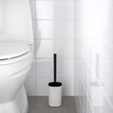 Escobilla de baño blanca en soporte negro junto a un inodoro en un baño minimalista.