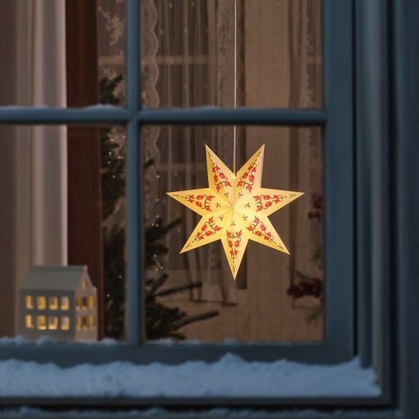 Lámpara de estrella escandinava en ventana con flores rojas, que brilla cálidamente, creando un ambiente interior acogedor con vista nevada.