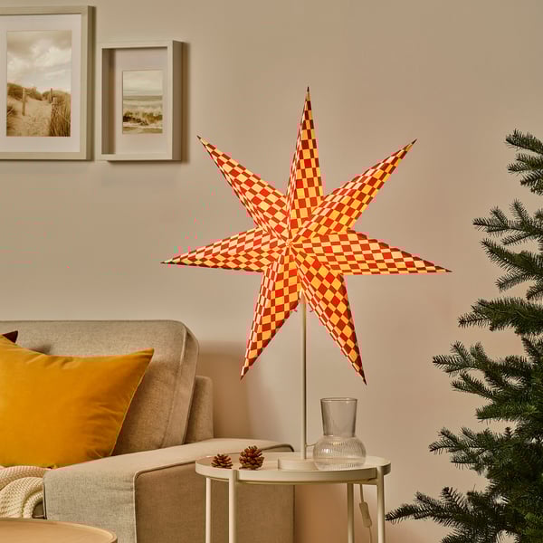 Linterna de estrella roja y amarilla STRÅLA cerca del árbol de Navidad, más alta que el sillón, sobre mesa blanca con piñas.