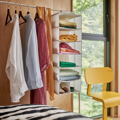 Una escena de dormitorio con una sábana a rayas, dos perchas que sostienen ropa y una silla amarilla junto a una ventana.