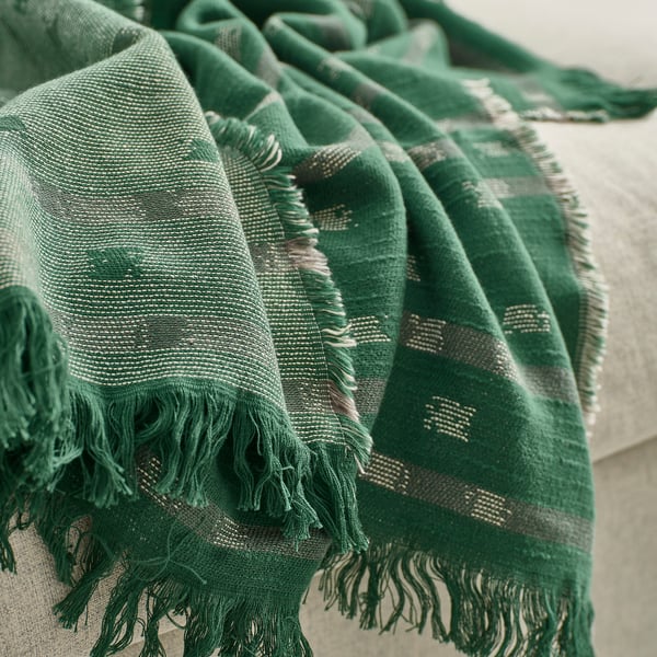 Manta de algodón tejido en jacquard verde oscuro con motivos y flecos en blanco roto/gris, drapeada sobre una superficie de color claro, que muestra textura y calidad.