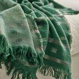 Manta de algodón tejido en jacquard verde oscuro con motivos y flecos en blanco roto/gris, drapeada sobre una superficie de color claro, que muestra textura y calidad.