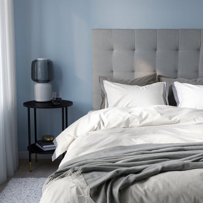 Cama azul moderna con edredón TÅTELSMYGARE blanco, manta gris, cabecero gris y mesita auxiliar negra.