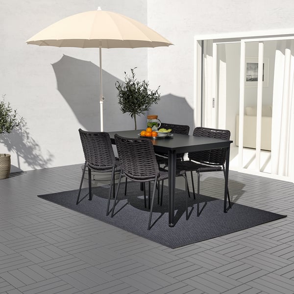 Patio al aire libre con sillas de mimbre negro y mesa rectangular, además de una sombrilla beige, dispuesta sobre una terraza gris para comer.