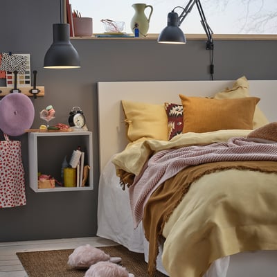 Dormitorio con cama acogedora, ropa de cama mostaza, estantería de pared con libros, lámpara, reloj y artículos decorativos.