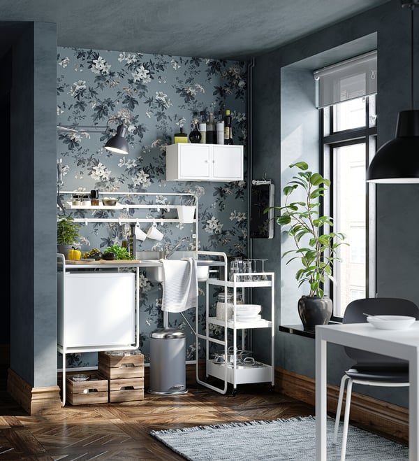 Cocina moderna con papel tapiz floral, nevera pequeña y carritos de almacenamiento. Decoración blanca y acentos negros, gran ventanal, que añade luminosidad.