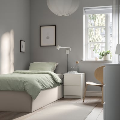 Dormitorio gris minimalista con cama TJÅLLE color menta, mesita de noche blanca, arte abstracto, lámpara colgante, silla de madera y plantas.