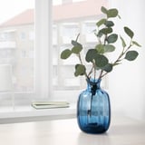 Jarrón de vidrio azul sobre mesa blanca cerca de la ventana con ramas de plantas verdes.