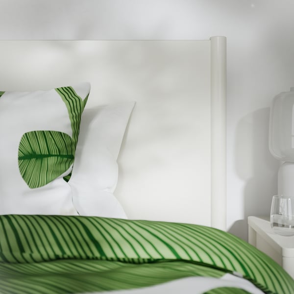 Cama blanca moderna con cajones de calidad, almohadas con patrón de hojas verdes, mesita de noche blanca cuidada, lámpara elegante.