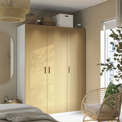 Dormitorio moderno con gran armario TONSTAD de madera clara, cama blanca, silla de mimbre y planta verde.