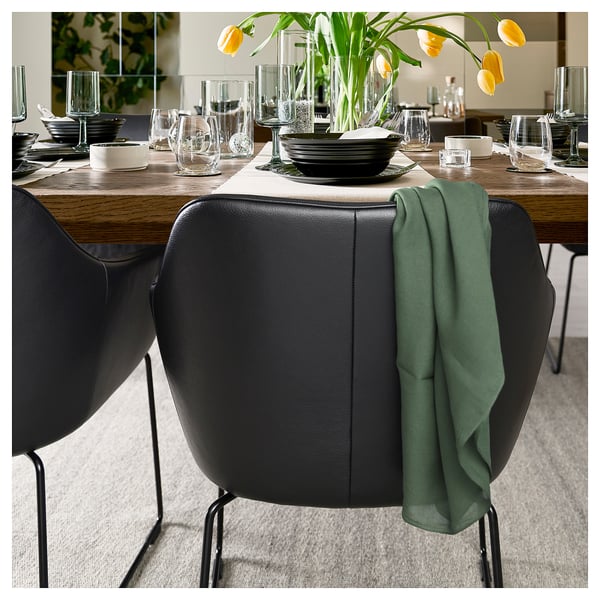 Elegante comedor con sillas de cuero negro TOSSBERG, mesa de madera, servilletas verdes, copas de vino y arreglos florales. Moderno y acogedor.