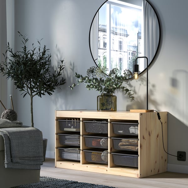 Afirmado, mueble de almacenaje de madera clara TROFAST, cajas de malla gris, espejo de pared redondo, planta verde, sofá gris, suelo de madera, pared gris claro.