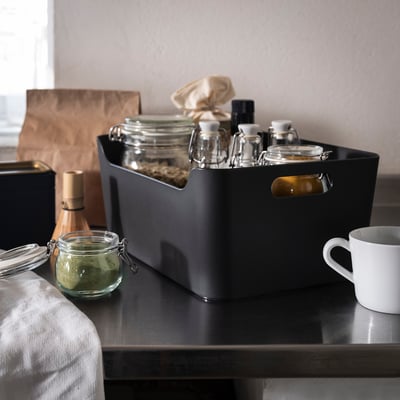 Caja UPPDATERA negra en mostrador con frascos y taza, encaja bien entre los artículos de cocina.
