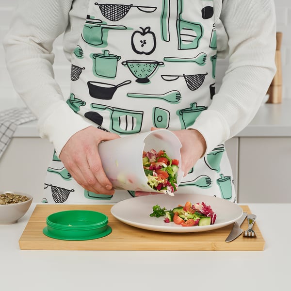 Persona sosteniendo la coctelera de ensaladas UPPFYLLD, vertiendo ensalada enjuagada en un plato sobre una tabla de madera. Los diseños de utensilios de cocina del delantal.