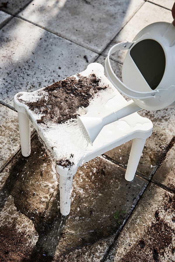 Taburete infantil de plástico blanco con bordes y patas redondeadas, parcialmente cubierto de tierra, junto a una regadera blanca sobre un suelo de piedra.
