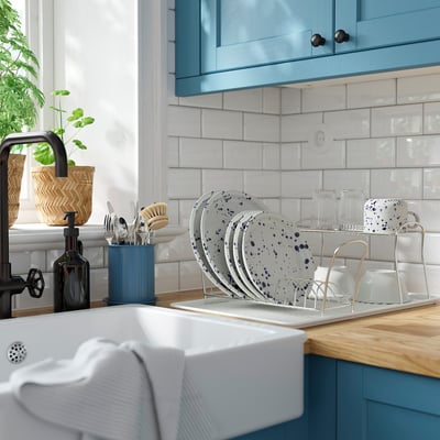 Fregaderos de cocina con escurridor para platos con 10 platos, cubiertos y tazas. Encimera de madera, armarios azules y salpicadero de baldosas.