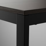 Primer plano de la mesa VANGSTA: elegante tablero de madera oscura con acabado liso se combina con robustas patas de metal negro. Diseñado para ser duradero.
