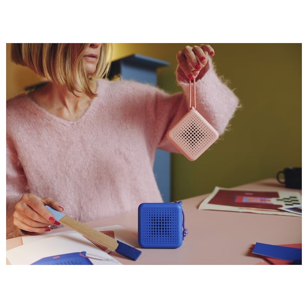 Una persona con un suéter rosa sostiene un altavoz portátil rosa mientras está sentada en una mesa con artículos de manualidades. Un altavoz azul también está sobre la mesa.