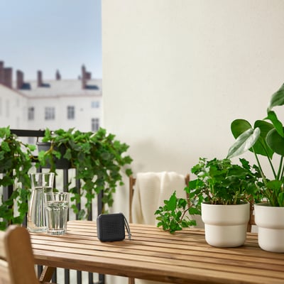 Pequeño altavoz VAPPEBY negro se encuentra sobre una mesa de madera entre plantas y vasos. Ambientación de balcón urbano.