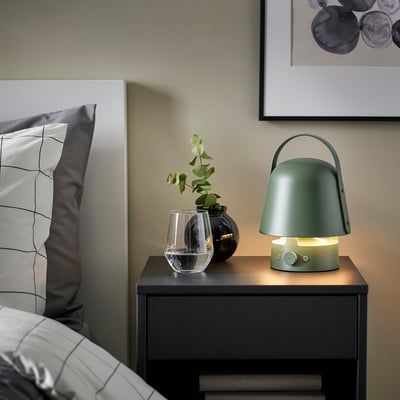 Lámpara VAPPEBY verde en mesita de noche, con planta y almohada. Portátil, brillo suave.