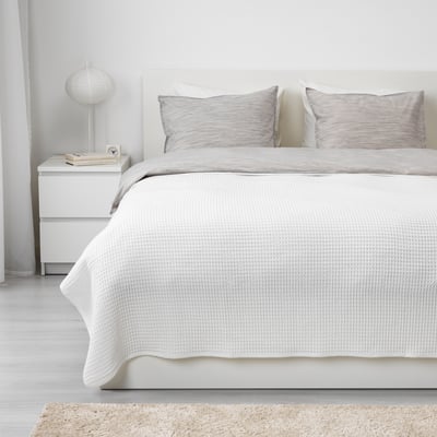 Colcha blanca sobre cama doble con almohadas grises, mesita de noche con lámpara, libro y foto.