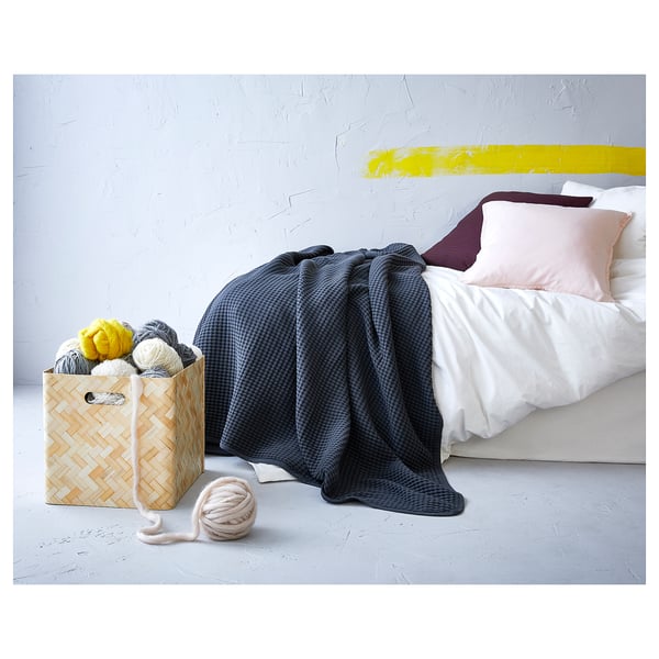 Una acogedora escena de dormitorio con una colcha VÅRELD de algodón tejido azul, almohadas rosas y moradas y una cesta con hilo de colores.