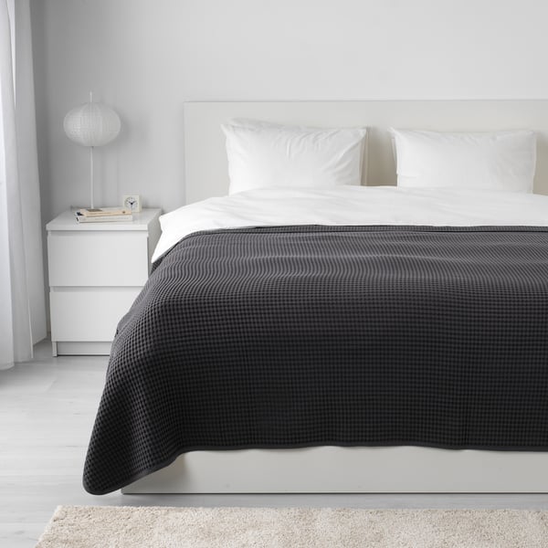 Dormitorio con cama blanca, colcha VÅRELD gris oscuro, almohadas blancas, mesita de noche, lámpara, reloj, libros, alfombra grande.