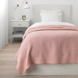 Acogedor dormitorio con colcha rosa claro, cabecero gris, mesita de noche y lámpara.