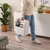Persona sosteniendo un organizador portátil VATTENKAR con computadora portátil, auriculares y archivos, de pie junto a gabinetes y plantas blancas.