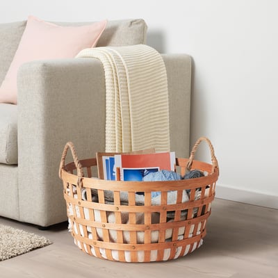 Cesta beige con asas de madera, cargadores y una manta, junto a un sofá gris.
