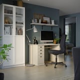 Una moderna configuración de oficina en casa con un escritorio blanco, una silla ergonómica gris y una estantería, dispuestos contra una pared verde.