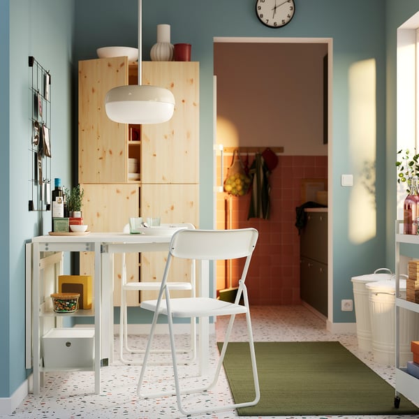 Cocina moderna con armarios de madera blanca y paredes azules. Silla plegable VIHALS blanca sobre mesa de pino. Diseño luminoso, espacioso y sencillo.