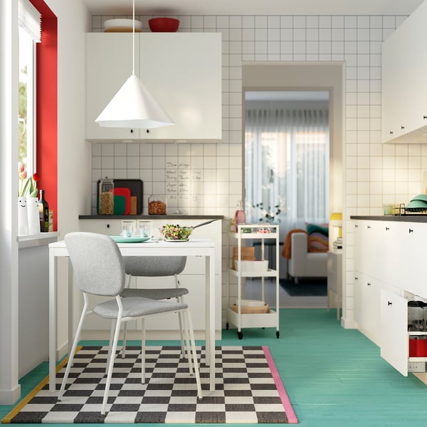 Cocina moderna con mesa VIHALS y silla gris, alfombra a cuadros, lámpara colgante blanca, suelo verde, sala de estar abierta.