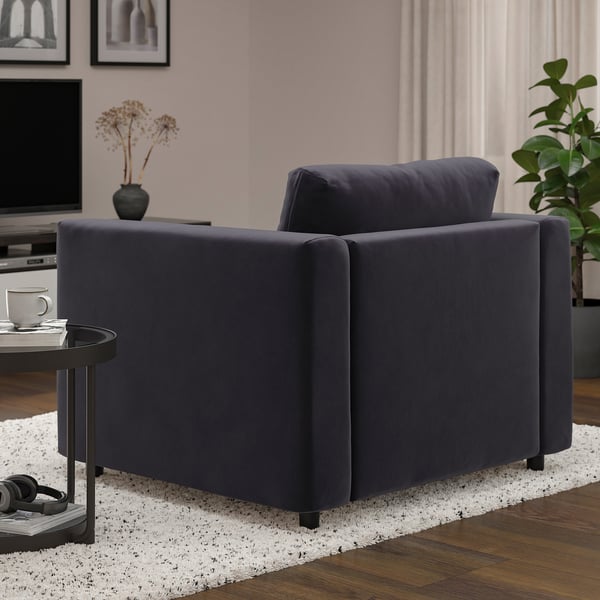 Un acogedor sillón gris oscuro en un espacio habitable moderno. A su lado, una pequeña mesa contiene una taza y auriculares. Detrás, un televisor y un jarrón con flores secas.