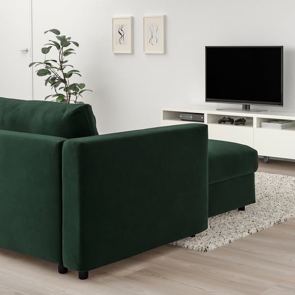 Sala de estar moderna con sofá seccional VIMLE verde esmeralda, piso de madera, mueble de TV blanco, planta y obras de arte.