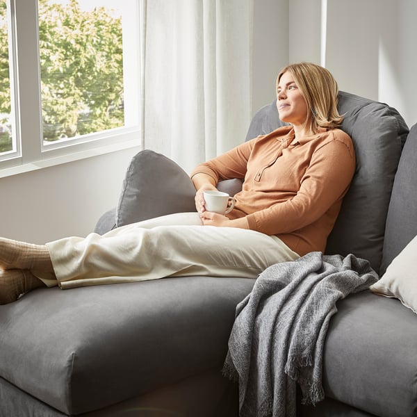 Mujer relajada en el sofá seccional gris VINLIDEN junto a la ventana, sosteniendo una taza de café, acogedora con manta beige.