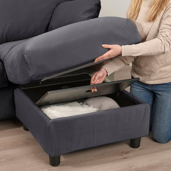 Una persona abre un sofá otomano gris para revelar un espacio de almacenamiento lleno de almohadas, mostrando su diseño funcional.