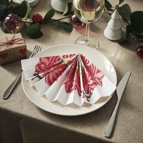 Una mesa puesta con platos blancos y cubiertos. Coloridas servilletas dobladas se colocan encima de cada plato. Copas de vino y decoraciones navideñas rodean el entorno.