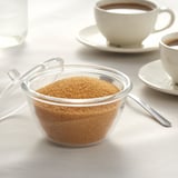 Recipiente de vidrio transparente con azúcar, cucharas y tazas de café sobre un mantel.
