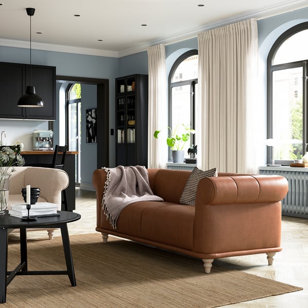 Sala de estar moderna con sofá de cuero marrón VISKAFORS, cortinas beige, ventanas arqueadas, mesa de centro negra y cocina de madera.